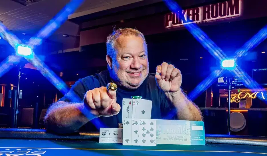 Kuvassa on riemukkaasti hymyilevä mies, joka poseeraa pokeriturnauksen voittajana. Hän istuu kirkkaansinisen pelipöydän ääressä ja ojentaa toista kättään ylpeänä eteenpäin, esitellen suurta kultaista mestaruussormusta sormessaan. Hänen edessään pöydällä on voittokäsi – neljä ässää – sekä ensimmäisen sijan palkintolaatta ja suuri "Ticket to Paradise" -voittokuponki. Taustalla näkyy "POKER ROOM" -kyltti, ja kuvaan luovat dramaattisen efektin kirkkaat siniset valot, jotka muodostavat ristikkäisen kuvion. Miehen ilme on täynnä aitoa iloa ja tyytyväisyyttä.