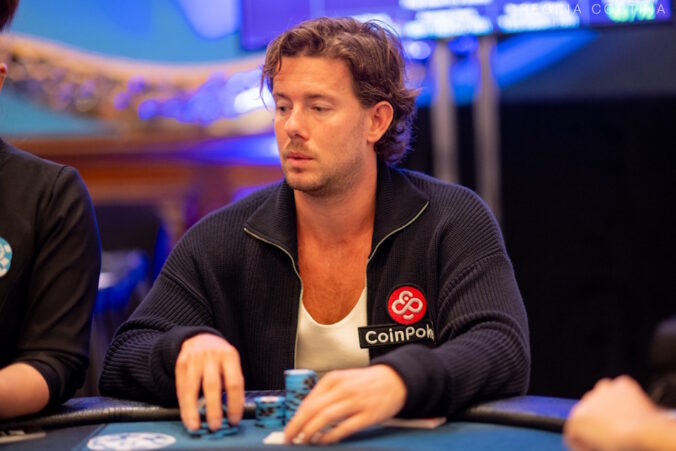 Mies istuu pokeripöydän ääressä syvän keskittyneenä ja analyyttinen ilme kasvoillaan. Mihellä on yllään tummansininen, vetoketjullinen neuletakki, jonka rinnassa on selkeästi näkyvä CoinPoker-pokerisivuston logo, mikä viittaa hänen sponsorisuhteeseensa. Kuva on otettu ammattilaisturnauksen aikana, ja taustan kirkkaat, epätarkat valot luovat peliin jännittyneen ja korkeatasoisen tunnelman. Gerritsen kädet ovat pöydällä pelimerkkien lähellä, valmiina tekemään siirtonsa.
