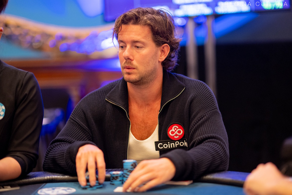 Mies istuu pokeripöydän ääressä syvän keskittyneenä ja analyyttinen ilme kasvoillaan. Mihellä on yllään tummansininen, vetoketjullinen neuletakki, jonka rinnassa on selkeästi näkyvä CoinPoker-pokerisivuston logo, mikä viittaa hänen sponsorisuhteeseensa. Kuva on otettu ammattilaisturnauksen aikana, ja taustan kirkkaat, epätarkat valot luovat peliin jännittyneen ja korkeatasoisen tunnelman. Gerritsen kädet ovat pöydällä pelimerkkien lähellä, valmiina tekemään siirtonsa.