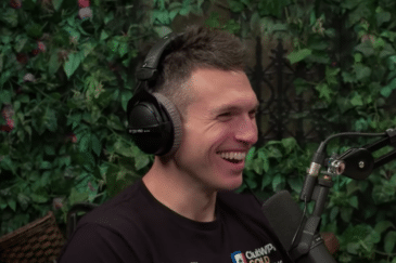 Kuvassa miehellä on musta paita ja mustat kuulokkeet. Hän hymyilee. Taustalla näkyy vihreitä puunlehtiä.