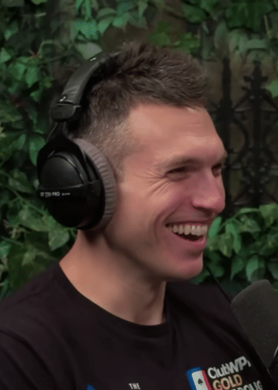 Kuvassa miehellä on musta paita ja mustat kuulokkeet. Hän hymyilee. Taustalla näkyy vihreitä puunlehtiä.