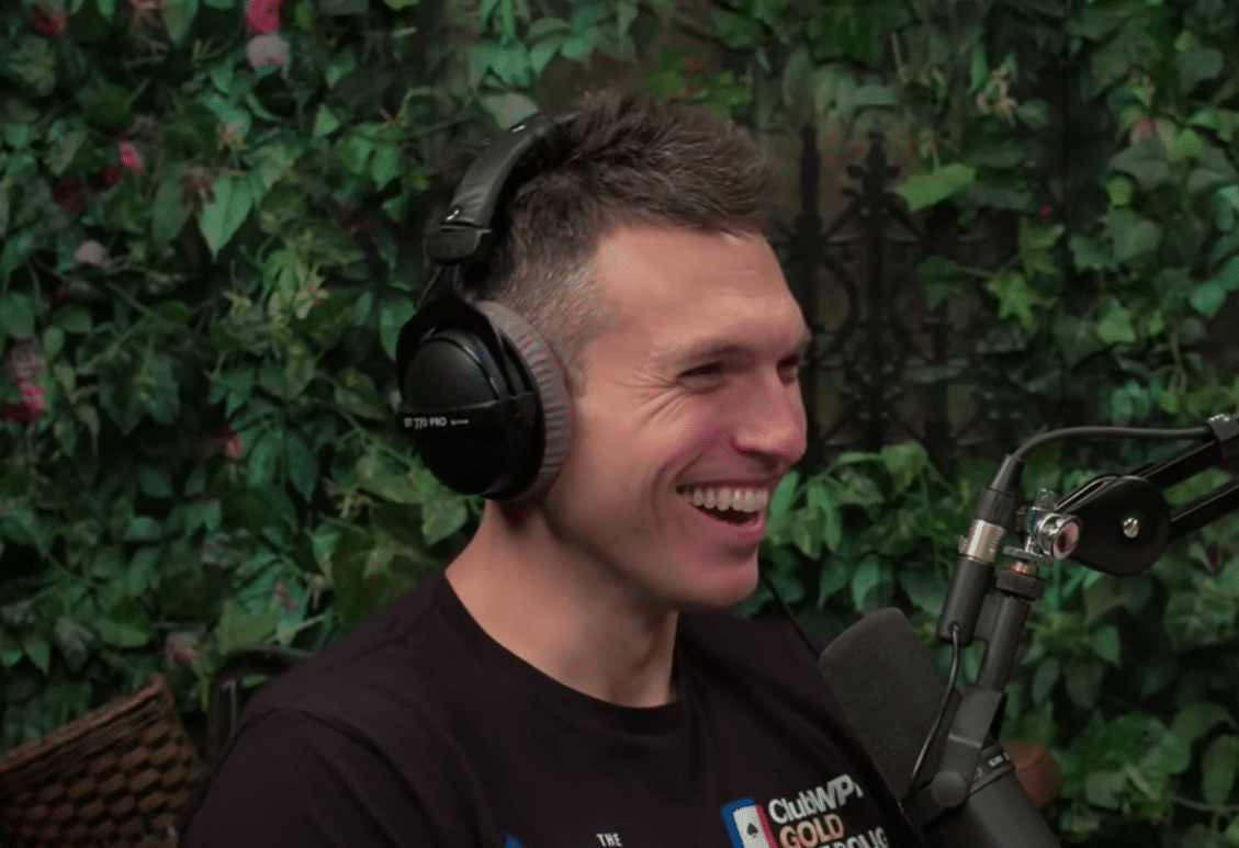 Kuvassa miehellä on musta paita ja mustat kuulokkeet. Hän hymyilee. Taustalla näkyy vihreitä puunlehtiä.