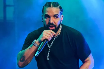 Kuvassa on maailmankuulu kanadalainen artisti Drake esiintymässä lavalla. Hän katsoo intensiivisesti suoraan kameraan samalla kun hän räppää mikrofoniin, jota hän pitelee toisessa kädessään. Hänen hiuksensa on letitetty cornrows-tyyliin, ja hänellä on yllään yksinkertainen musta t-paita. Draken olemusta korostavat lukuisat näyttävät korut, kuten suuri timanttikaulaketju, kello ja useita rannekoruja, sekä hänen käsivarsissaan näkyvät tatuoinnit. Tunnelmallinen, voimakkaan sininen ja viileä lavavalaistus ympäröi hänet, luoden dynaamisen ja keskittyneen tunnelman kesken live-esiintymisen.