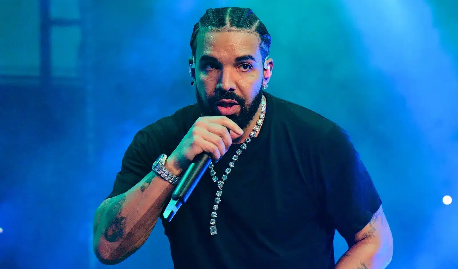 Kuvassa on maailmankuulu kanadalainen artisti Drake esiintymässä lavalla. Hän katsoo intensiivisesti suoraan kameraan samalla kun hän räppää mikrofoniin, jota hän pitelee toisessa kädessään. Hänen hiuksensa on letitetty cornrows-tyyliin, ja hänellä on yllään yksinkertainen musta t-paita. Draken olemusta korostavat lukuisat näyttävät korut, kuten suuri timanttikaulaketju, kello ja useita rannekoruja, sekä hänen käsivarsissaan näkyvät tatuoinnit. Tunnelmallinen, voimakkaan sininen ja viileä lavavalaistus ympäröi hänet, luoden dynaamisen ja keskittyneen tunnelman kesken live-esiintymisen.