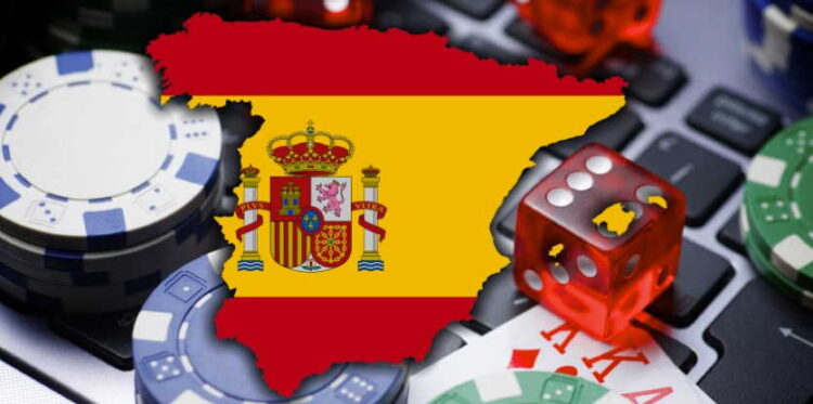 Kuvassa on digitaalinen sommitelma, joka kuvaa online-rahapelaamista Espanjassa. Kuvan keskeisenä elementtinä on Espanjan kartan muotoinen kuvio, joka on täytetty maan lipun väreillä ja vaakunalla. Tämä on asetettu kannettavan tietokoneen näppäimistön päälle, mikä symboloi internetiä ja verkkoympäristöä. Ympärillä on uhkapelaamiseen liittyviä esineitä, kuten pokerimerkkejä, läpikuultava punainen noppa ja pelikortteja (ässä ja kuningas). Kokonaisuus on tyypillinen kuvituskuva, joka viestii tehokkaasti Espanjan online-pelimarkkinoista, lainsäädännöstä tai maan pelikulttuurista.
