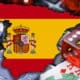 Kuvassa on digitaalinen sommitelma, joka kuvaa online-rahapelaamista Espanjassa. Kuvan keskeisenä elementtinä on Espanjan kartan muotoinen kuvio, joka on täytetty maan lipun väreillä ja vaakunalla. Tämä on asetettu kannettavan tietokoneen näppäimistön päälle, mikä symboloi internetiä ja verkkoympäristöä. Ympärillä on uhkapelaamiseen liittyviä esineitä, kuten pokerimerkkejä, läpikuultava punainen noppa ja pelikortteja (ässä ja kuningas). Kokonaisuus on tyypillinen kuvituskuva, joka viestii tehokkaasti Espanjan online-pelimarkkinoista, lainsäädännöstä tai maan pelikulttuurista.