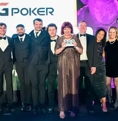 Kuvassa on yhdeksän hengen ryhmä, joka poseeraa juhlallisesti lavalla, todennäköisesti palkintotilaisuudessa. Taustalla näkyy suuri näyttö, jossa on GGPoker-logo, sekä näyttävä, violetilla valaistu patsas. Ryhmän miehet ovat pukeutuneet pääasiassa tummiin pukuihin ja smokkeihin, ja naiset tyylikkäisiin ilta-asuihin. Keskellä seisova nainen pitelee ylpeänä käsissään palkintolaattaa, ja koko ryhmä hymyileekin iloisesti kameralle. Valaistus on ammattimainen ja tunnelma on selvästi juhlava ja onnistunut, vangiten tärkeän palkitsemishetken.