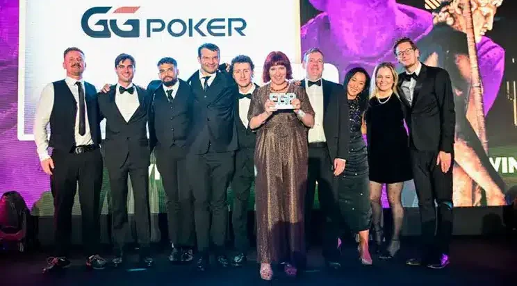 Kuvassa on yhdeksän hengen ryhmä, joka poseeraa juhlallisesti lavalla, todennäköisesti palkintotilaisuudessa. Taustalla näkyy suuri näyttö, jossa on GGPoker-logo, sekä näyttävä, violetilla valaistu patsas. Ryhmän miehet ovat pukeutuneet pääasiassa tummiin pukuihin ja smokkeihin, ja naiset tyylikkäisiin ilta-asuihin. Keskellä seisova nainen pitelee ylpeänä käsissään palkintolaattaa, ja koko ryhmä hymyileekin iloisesti kameralle. Valaistus on ammattimainen ja tunnelma on selvästi juhlava ja onnistunut, vangiten tärkeän palkitsemishetken.