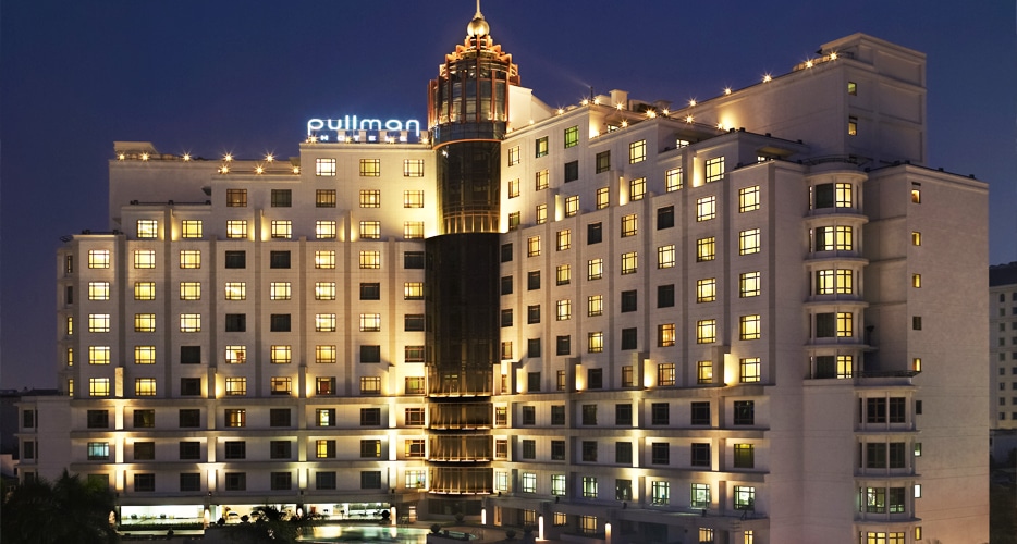 Kuvassa on suuri ja ylellisen näköinen Pullman-hotellirakennus, joka on kuvattu iltahämärässä. Rakennuksen pääosin valkoinen, monitasoinen julkisivu on upeasti valaistu. Sen lukuisat neliönmuotoiset ikkunat hohtavat lämmintä keltaista valoa pimeää, syvänsinistä taivasta vasten. Arkkitehtuurin keskeinen elementti on moderni, lasinen ja pyöreä torni, joka kohoaa rakennuksen keskeltä ja jonka kruunaa koristeellinen huippu. Aivan katon rajassa loistaa brändin tunnusomainen sininen 'pullman'-valomainos. Kuva luo hienostuneen ja kutsuvan tunnelman.