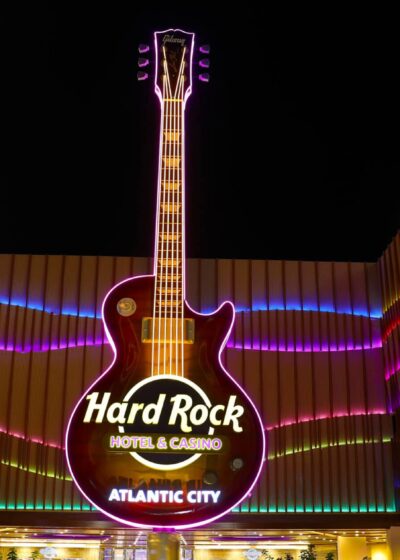 Kuvassa on Atlantic Cityssä sijaitsevan Hard Rock Hotel & Casinon näyttävä sisäänkäynti upeassa yövalaistuksessaan. Kuvan absoluuttinen keskipiste ja katseenvangitsija on valtava, pystysuora sähkökitara, joka toimii rakennuksen ikonisena neonkylttinä. Kitaran rungossa lukee "Hard Rock HOTEL & CASINO ATLANTIC CITY". Moderni rakennus reunustaa kitaraa molemmin puolin, ja sen julkisivua koristavat dynaamiset ja värikkäät, aaltoilevat valot – pääasiassa pinkin, purppuran ja vihreän sävyissä. Valaistu katos peittää ajoväylän, luoden ylellisen ja energisen tunnelman tälle kuuluisalle viihdekeskukselle.