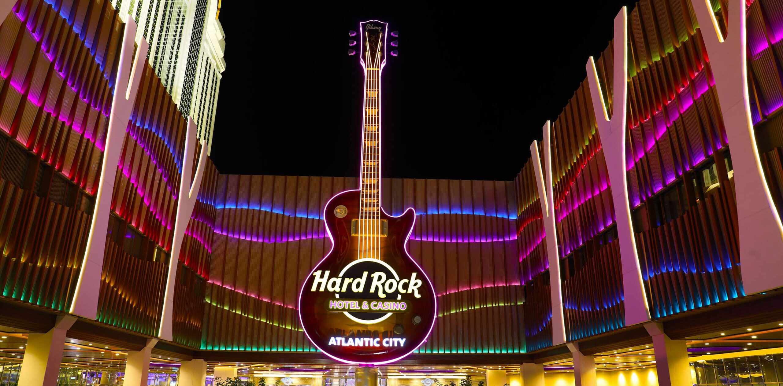 Kuvassa on Atlantic Cityssä sijaitsevan Hard Rock Hotel & Casinon näyttävä sisäänkäynti upeassa yövalaistuksessaan. Kuvan absoluuttinen keskipiste ja katseenvangitsija on valtava, pystysuora sähkökitara, joka toimii rakennuksen ikonisena neonkylttinä. Kitaran rungossa lukee "Hard Rock HOTEL & CASINO ATLANTIC CITY". Moderni rakennus reunustaa kitaraa molemmin puolin, ja sen julkisivua koristavat dynaamiset ja värikkäät, aaltoilevat valot – pääasiassa pinkin, purppuran ja vihreän sävyissä. Valaistu katos peittää ajoväylän, luoden ylellisen ja energisen tunnelman tälle kuuluisalle viihdekeskukselle.