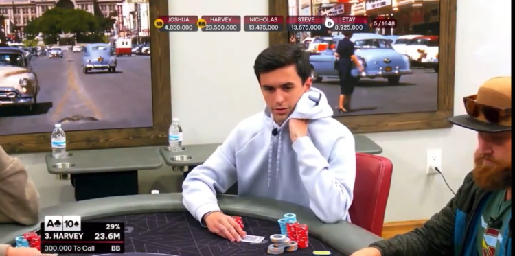 Kuvassa on nuori pokerinpelaaja nimeltä Harvey, joka on keskittynyt tiukasti käynnissä olevaan pokerikäteen. Kuva on peräisin televisioidusta tai suoratoistetusta pelistä. Harvey, pukeutuneena vaaleanharmaaseen huppariin, istuu mietteliäänä ja nojaa poskeaan käteensä. Kuvan vasemmassa alareunassa oleva grafiikka paljastaa hänen kätensä olevan pata-ässä ja patakymppi (A♠ 10♠) ja hänen voittotodennäköisyytensä olevan 29%. Grafiikka näyttää myös, että hänen pitäisi maksaa 300 000 jatkaakseen pelissä. Taustalla on mielenkiintoisena yksityiskohtana suuria, kehystettyjä kuvia vanhoista 1950-luvun autoista katunäkymässä. Tunnelma pöydässä on jännittynyt ja keskittynyt.