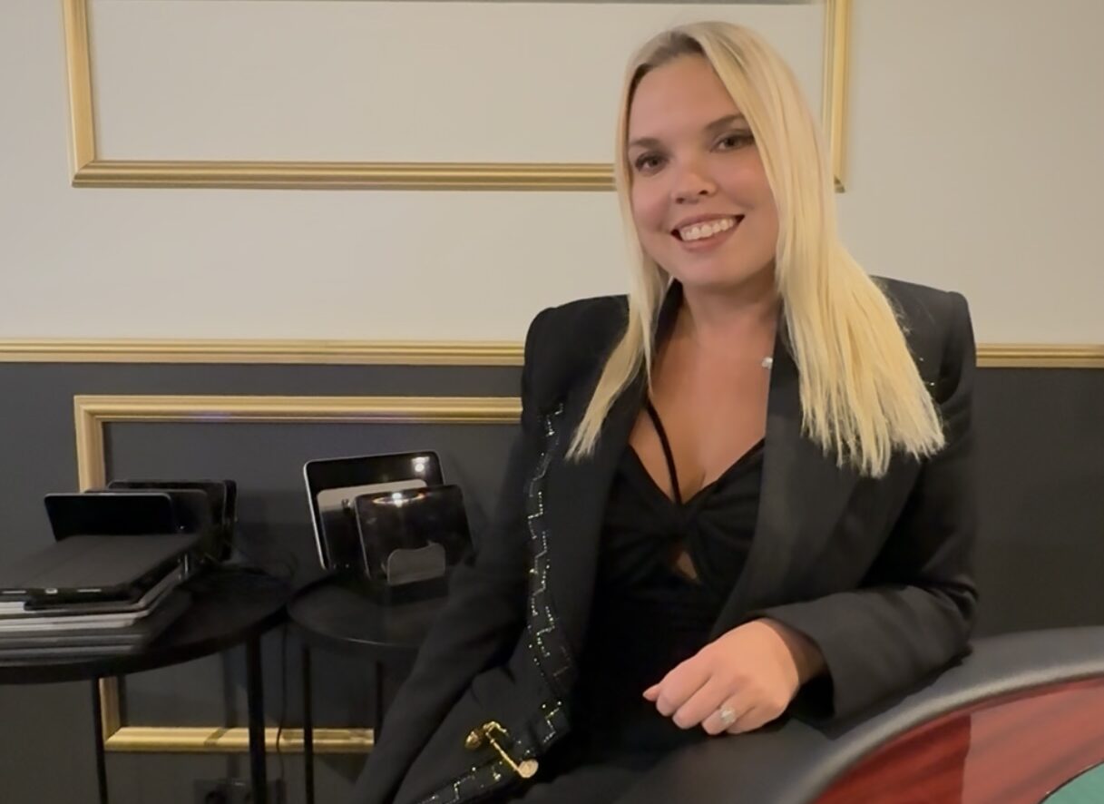 Kerli Kyllönen istuu pokeripöydän ääressä mustassa topissa ja jakussa. Taustalla pöytää ja valokuvataidetta