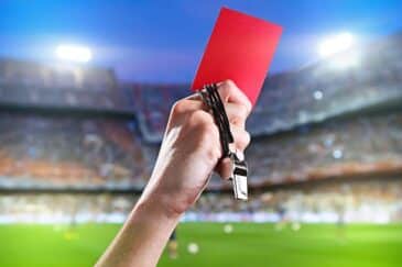 Kuvassa on jalkapallotuomarin käsi, joka on kohotettu määrätietoisesti ilmaan. Kädessä on punainen kortti, joka on vakavin rangaistus jalkapallossa ja tarkoittaa pelaajan poistamista kentältä. Tuomarin sormien ympärille on kietoutunut myös musta kaulanauha, jossa roikkuu metallinen pilli. Toiminta tapahtuu suuren ja kirkkaasti valaistun jalkapallostadionin vihreällä nurmella. Taustalla näkyvät epätarkat katsomot ovat täynnä yleisöä, mikä luo kuvaan dramaattisen ja tärkeän ottelun tunnelman.i