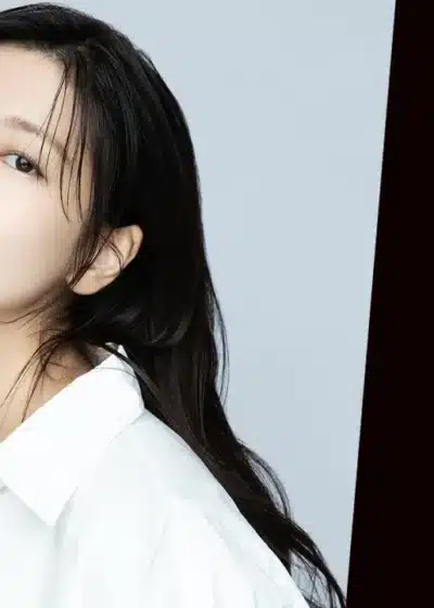 Kuvassa on mainosmainen sommitelma, joka on jaettu kahteen osaan. Vasemmalla puolella on nuori, itäaasialainen nainen, jolla on pitkät, tummat hiukset ja yllään valkoinen kauluspaita. Hän katsoo kameraan lempeällä ilmeellä, toinen käsi kevyesti kohotettuna poskensa viereen. Hänen olemuksensa on raikas ja luonnollinen. Oikealla puolella on mustalla taustalla suuri, kirkkaanpunainen Netflix-logo. Asetelma viittaa vahvasti siihen, että kuvassa oleva nainen on näyttelijä, joka tähdittää jotakin Netflix-tuotantoa.