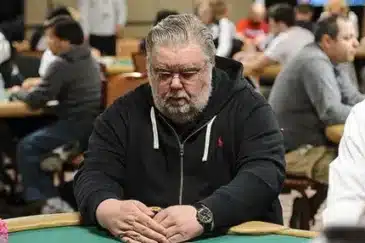 mies istuu pokeripöydässsä. Hänellä on päällään tumma huppari. Miehellä on kädet pöydällä ja hän peittää pelikortit.