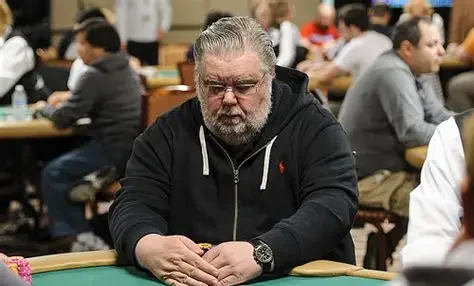 mies istuu pokeripöydässsä. Hänellä on päällään tumma huppari. Miehellä on kädet pöydällä ja hän peittää pelikortit.