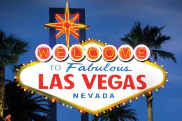 Kuvassa on maailmankuulu "Tervetuloa to Fabulous Las Vegas, Nevada" -kyltti, joka on kuvattu ikonisessa iltavalaistuksessaan. Taivas on syvän sininen hämärän hetkellä, mikä luo täydellisen taustan kirkkaasti hohtavalle kyltille. Kyltti on vinoneliön muotoinen, ja sen yläosassa loistaa suuri punakeltainen neon-tähti. Sana "WELCOME" on kirjoitettu yksittäisiin, hehkuviin ympyröihin. Tämän alapuolella on teksti "to Fabulous" sinisellä ja "LAS VEGAS" suurilla punaisilla kirjaimilla. Kyltin molemmin puolin kohoaa tummia palmujen siluetteja, jotka viimeistelevät tämän klassisen ja kutsuvan näkymän.