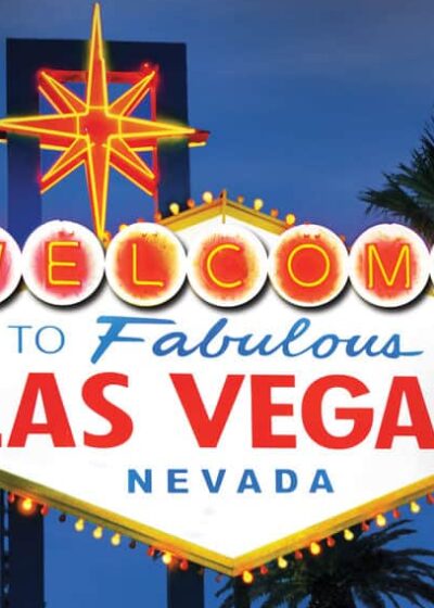 Kuvassa on maailmankuulu "Tervetuloa to Fabulous Las Vegas, Nevada" -kyltti, joka on kuvattu ikonisessa iltavalaistuksessaan. Taivas on syvän sininen hämärän hetkellä, mikä luo täydellisen taustan kirkkaasti hohtavalle kyltille. Kyltti on vinoneliön muotoinen, ja sen yläosassa loistaa suuri punakeltainen neon-tähti. Sana "WELCOME" on kirjoitettu yksittäisiin, hehkuviin ympyröihin. Tämän alapuolella on teksti "to Fabulous" sinisellä ja "LAS VEGAS" suurilla punaisilla kirjaimilla. Kyltin molemmin puolin kohoaa tummia palmujen siluetteja, jotka viimeistelevät tämän klassisen ja kutsuvan näkymän.