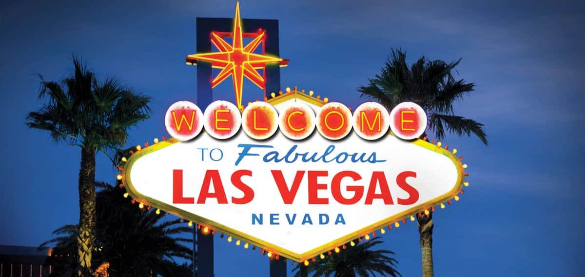 Kuvassa on maailmankuulu "Tervetuloa to Fabulous Las Vegas, Nevada" -kyltti, joka on kuvattu ikonisessa iltavalaistuksessaan. Taivas on syvän sininen hämärän hetkellä, mikä luo täydellisen taustan kirkkaasti hohtavalle kyltille. Kyltti on vinoneliön muotoinen, ja sen yläosassa loistaa suuri punakeltainen neon-tähti. Sana "WELCOME" on kirjoitettu yksittäisiin, hehkuviin ympyröihin. Tämän alapuolella on teksti "to Fabulous" sinisellä ja "LAS VEGAS" suurilla punaisilla kirjaimilla. Kyltin molemmin puolin kohoaa tummia palmujen siluetteja, jotka viimeistelevät tämän klassisen ja kutsuvan näkymän.