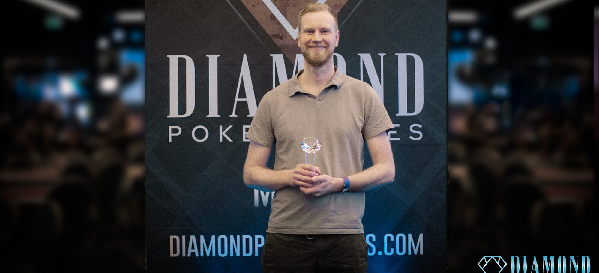 Mies seisoo palkinto kädessään. Hän hymyilee ja hänellä on päällään vaaleanruskea t-paita.Takana näkyy Diamond Poker Serieksen kyltti.