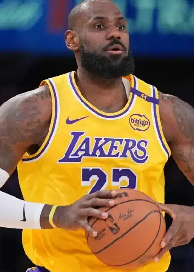 Kuvassa on yksi kaikkien aikojen tunnetuimmista koripalloilijoista, LeBron James. Hän seisoo kentällä pukeutuneena Los Angeles Lakersin ikoniseen kultaiseen pelipaitaan, jossa on purppuranväriset numerot 23 ja teksti "LAKERS". James pitää koripalloa molemmin käsin rintansa edessä ja katsoo keskittyneesti kentän sivuun. Hänen ilmeensä on vakava ja analyyttinen, kuin hän arvioisi pelitilannetta. Jamesilla on yllään myös valkoinen kompressiohiha vasemmassa kädessään, ja hänen tatuoidut kätensä näkyvät selvästi. Tausta on tumma ja sumea, korostaen häntä pelin polttopisteenä.