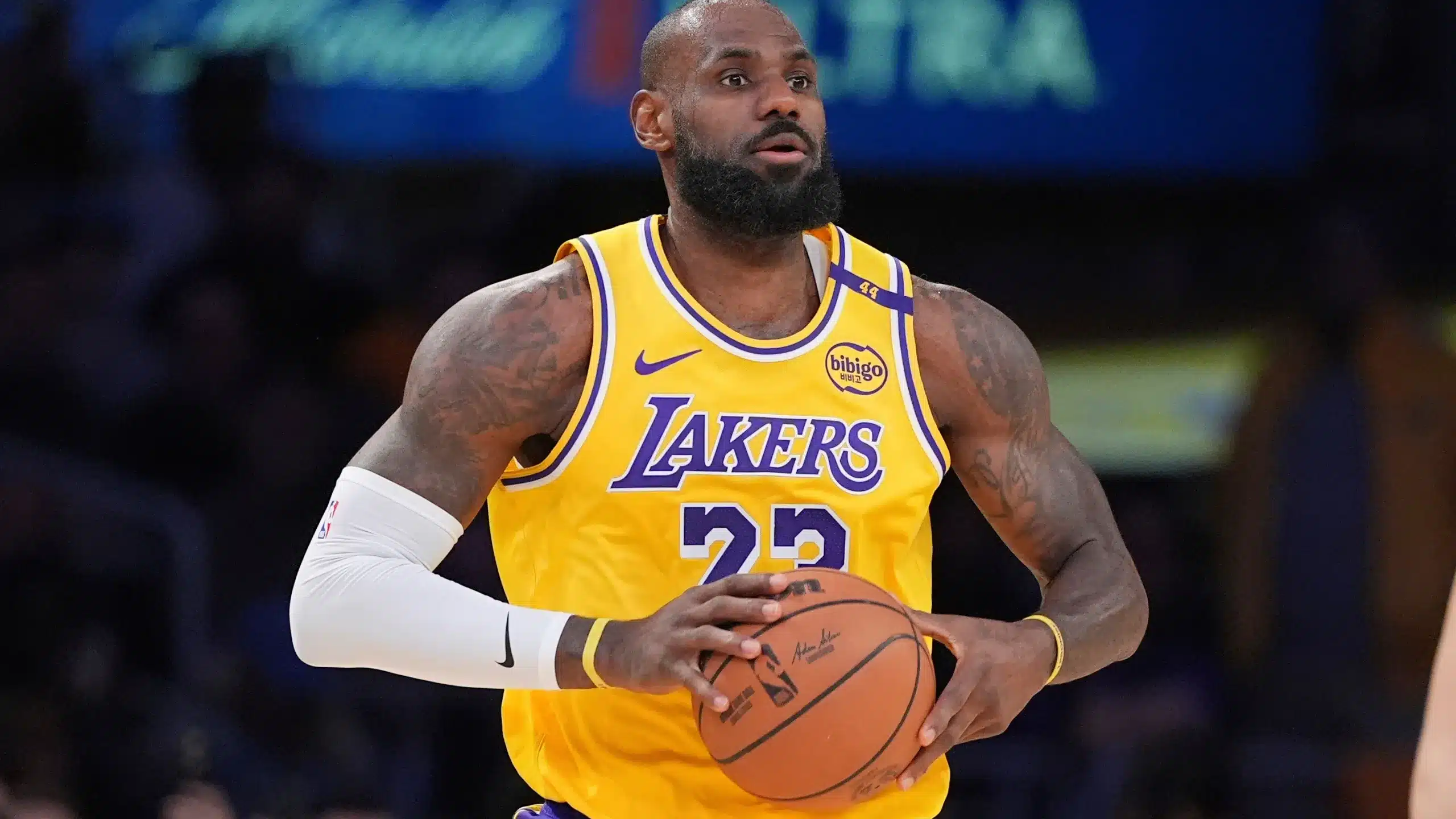 Kuvassa on yksi kaikkien aikojen tunnetuimmista koripalloilijoista, LeBron James. Hän seisoo kentällä pukeutuneena Los Angeles Lakersin ikoniseen kultaiseen pelipaitaan, jossa on purppuranväriset numerot 23 ja teksti "LAKERS". James pitää koripalloa molemmin käsin rintansa edessä ja katsoo keskittyneesti kentän sivuun. Hänen ilmeensä on vakava ja analyyttinen, kuin hän arvioisi pelitilannetta. Jamesilla on yllään myös valkoinen kompressiohiha vasemmassa kädessään, ja hänen tatuoidut kätensä näkyvät selvästi. Tausta on tumma ja sumea, korostaen häntä pelin polttopisteenä.