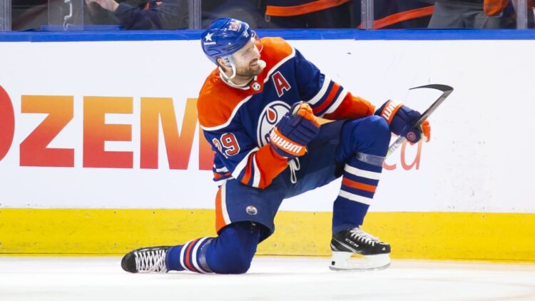 Kuvassa on saksalainen jääkiekkotähti Leon Draisaitl, joka pelaa NHL-joukkue Edmonton Oilersissa. Hän on polvistuneena yhdelle polvelle kesken ottelun, todennäköisesti tuulettamassa tekemäänsä maalia. Tämä on yksi hänen tunnetuimmista maalituuletuksistaan. Draisaitlilla on yllään Oilersin perinteinen sinioranssi peliasu, ja hänen rinnassaan on varakapteenin "A"-kirjain. Hänen pelinumeronsa on 29. Kuva on otettu NHL-ottelun aikana, ja taustalla näkyy kaukalon laita. Draisaitlin asento on voimakas ja dynaaminen, ja se vangitsee täydellisesti huippu-urheilun intensiteetin ja maalinteon riemun.