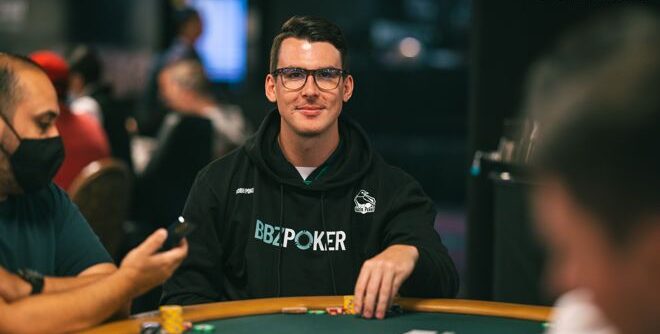 Kuvassa on nuorehko, silmälasipäinen mies, joka hymyilee ystävällisesti kameralle pokeriturnauksen aikana. Hän istuu vihreän pelipöydän ääressä ja on pukeutunut mustaan huppariin, jossa on näkyvä "BBZ POKER" -logo. Miehen käsi on pelimerkkien lähellä, mikä viittaa hänen olevan aktiivisesti mukana pelissä. Kuvan tausta on sumea, mutta siellä on erotettavissa kasinon valoja ja muita ihmisiä, mikä luo autenttisen turnaustunnelman. Kuvan oikeassa yläkulmassa on valokuvaaja Alec Romen vesileima.