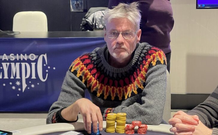 Mies istuu pokeripöydässä. Hänellä on päällään villapaita, jonka pohjaväri on harmaa. Paidassa on keltaisia, punaisia ja mustia kuvioita.