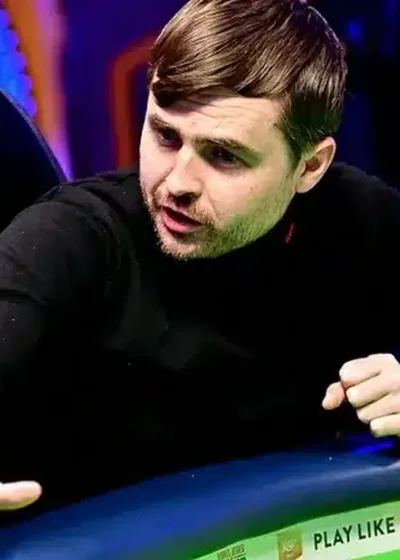 Kuvassa on intensiivinen hetki pokeriturnauksen pelipöydästä. Mustaan poolopaitaan pukeutunut pelaaja nojaa eteenpäin ja näyttää reagoivan voimakkaasti pelin tapahtumiin, joko puhumalla tai elehtimällä. Hänen kätensä on ojennettuna pöydän ylle, mikä lisää hetken dynaamisuutta. Pelaajan ilme on erittäin keskittynyt ja latautunut. Pöydällä hänen edessään on pinoja sinisiä ja keltaisia pelimerkkejä. Taustan epätarkat valot luovat ammattimaisen ja jännittyneen tunnelman. Valokuvaaja Tomáš Stacha on vanginnut hetken, joka on täynnä pokerin psykologista draamaa.