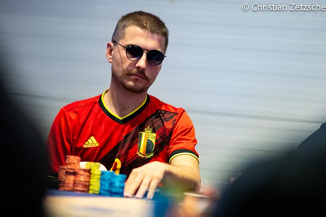Kuvassa on nuori mies keskittyneenä pokeripeliin viileä ja tarkkaavainen ilme kasvoillaan. Hänen persoonalliseen tyyliinsä kuuluvat lyhyet hiukset, näyttävät viikset sekä pyöreät aurinkolasit, jotka kätkevät hänen katseensa. Pelaajalla on yllään Belgian jalkapallomaajoukkueen punamusta pelipaita. Hänen kätensä on suurten ja värikkäiden pelimerkkipinojen äärellä, ja hän näyttää olevan keskellä peliä. Kuvan tunnelma on tyylikäs ja intensiivinen, ja sen on ottanut valokuvaaja Christian Zetzsche.