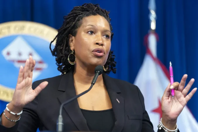Kuvassa on Washington D.C:n pormestari Muriel Bowser puhumassa lehdistötilaisuudessa. Hän seisoo puhujakorokkeella mikrofonin edessä ja elehtii käsillään ilmeikkäästi korostaakseen viestiään, pitäen toisessa kädessään vaaleanpunaista kynää. Hänellä on yllään musta jakku, ja hänen hiuksensa ovat rastoilla. Bowserin ilme on vakava ja keskittynyt. Taustalla näkyy osittain Washington D.C:n lippu sekä sininen taustakangas, jossa on kaupungin sinetti. Kuva välittää virallisen ja ammattimaisen tunnelman, jossa johtaja kommunikoi tärkeää asiaa.