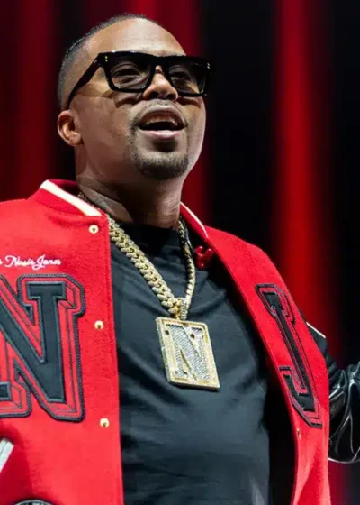 Kuvassa on yhdysvaltalainen rap-legenda Nas, joka on täydessä esiintymisvauhdissa lavalla. Hänellä on yllään näyttävä puna-musta college-takki, jossa on suuri "N"-kirjain, sekä musta t-paita ja tummat aurinkolasit. Hänen kaulassaan roikkuu massiivinen kultaketju, jossa on suuri N-riipus. Nas pitää toisessa kädessään mikrofonia ja osoittaa sormellaan ilmaan, samalla kun toinen käsi on koholla elehtien yleisölle. Hänen ilmeensä on energinen ja keskittynyt, vangiten täydellisesti live-esiintymisen intensiivisen tunnelman punaisen lavavalaistuksen keskellä.