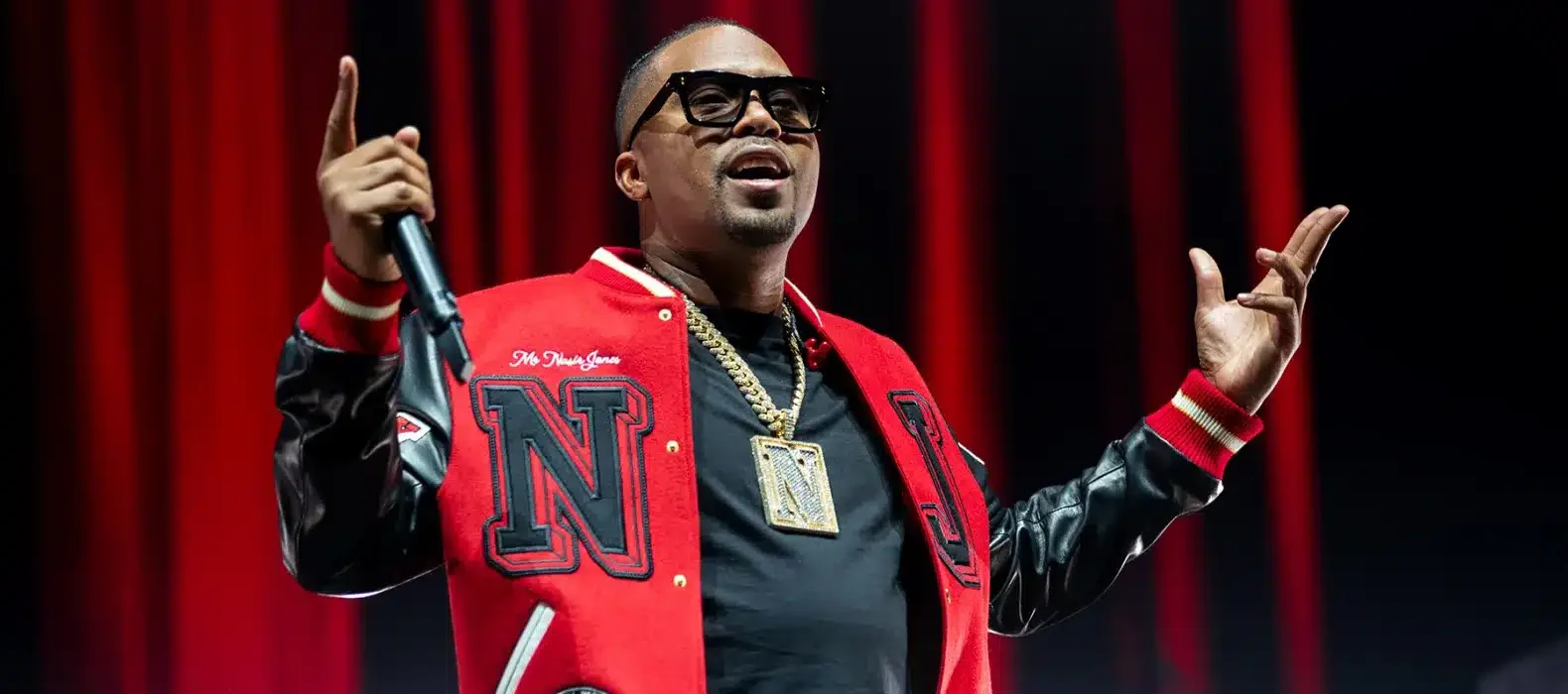Kuvassa on yhdysvaltalainen rap-legenda Nas, joka on täydessä esiintymisvauhdissa lavalla. Hänellä on yllään näyttävä puna-musta college-takki, jossa on suuri "N"-kirjain, sekä musta t-paita ja tummat aurinkolasit. Hänen kaulassaan roikkuu massiivinen kultaketju, jossa on suuri N-riipus. Nas pitää toisessa kädessään mikrofonia ja osoittaa sormellaan ilmaan, samalla kun toinen käsi on koholla elehtien yleisölle. Hänen ilmeensä on energinen ja keskittynyt, vangiten täydellisesti live-esiintymisen intensiivisen tunnelman punaisen lavavalaistuksen keskellä.