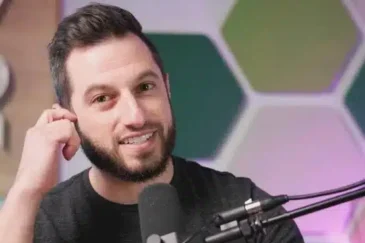 Kuvassa tummahiuksinen mies koskee korvaansa ja hänen edessään on mikrofoni. Tausta on värikäs.