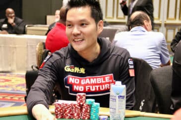 Kuvassa on kuuluisa i'pokeriammattilainen Ren Lin, joka hymyilee rennosti kesken turnauspelin. Hän istuu vihreän pelipöydän ääressä pukeutuneena mustaan huppariin, jossa on näkyvä GGPoker-logo ja suuri teksti "NO GAMBLE, NO FUTURE". Juandan edessä pöydällä on siistejä pinoja punaisia ja sinisiä pelimerkkejä sekä juomapakkaus. Hänen ilmeensä on hyväntuulinen, ja taustalla näkyy epätarkkana suuri ja tyylikäs turnaussali. Kuva vangitsee hienosti yhden pokerin menestyneimmistä pelaajista nauttimassa pelistä