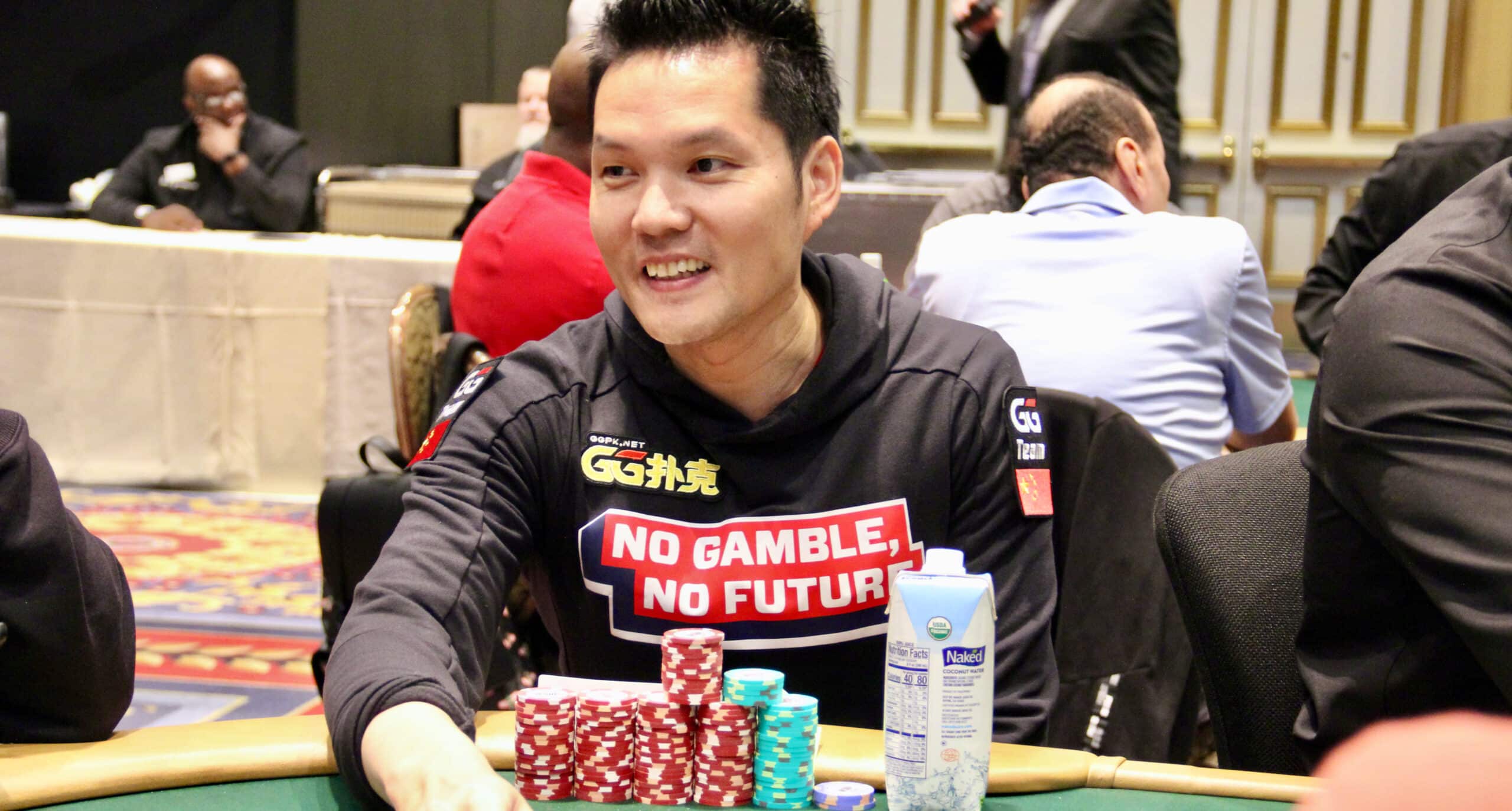 Kuvassa on kuuluisa i'pokeriammattilainen Ren Lin, joka hymyilee rennosti kesken turnauspelin. Hän istuu vihreän pelipöydän ääressä pukeutuneena mustaan huppariin, jossa on näkyvä GGPoker-logo ja suuri teksti "NO GAMBLE, NO FUTURE". Juandan edessä pöydällä on siistejä pinoja punaisia ja sinisiä pelimerkkejä sekä juomapakkaus. Hänen ilmeensä on hyväntuulinen, ja taustalla näkyy epätarkkana suuri ja tyylikäs turnaussali. Kuva vangitsee hienosti yhden pokerin menestyneimmistä pelaajista nauttimassa pelistä