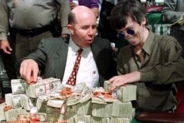 Kuvassa on ikoninen ja historiallinen hetki pokerimaailmasta. Siinä nähdään pokerilegenda Stu "The Kid" Ungar (oikealla) hänen voitettuaan World Series of Poker -pääturnauksen, todennäköisesti vuonna 1997. Ungar, jolla on tunnusomaiset pyöreät aurinkolasit, hymyilee leveästi. Hänen vieressään kasinojohtaja Jack Binion esittelee vaikuttavaa näkyä: vihreä pelipöytä on peitetty valtavalla määrällä käteistä rahaa, joka on pinottu korkeiksi nupuiksi, sekä tuhansilla pokerimerkeillä. Tämä oli perinteinen tapa esitellä turnauksen miljoonan dollarin pääpalkintoa. Kuva vangitsee täydellisesti vanhan ajan Las Vegasin tunnelman ja valtavan voiton suuruuden.