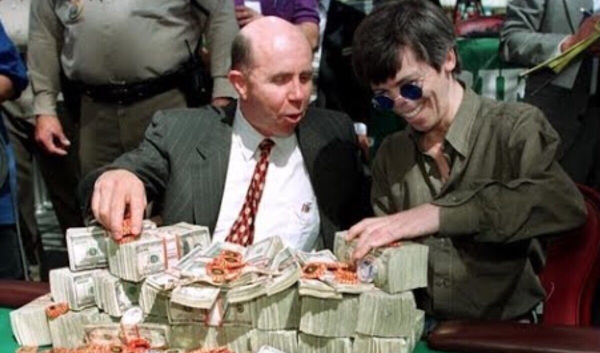 Kuvassa on ikoninen ja historiallinen hetki pokerimaailmasta. Siinä nähdään pokerilegenda Stu "The Kid" Ungar (oikealla) hänen voitettuaan World Series of Poker -pääturnauksen, todennäköisesti vuonna 1997. Ungar, jolla on tunnusomaiset pyöreät aurinkolasit, hymyilee leveästi. Hänen vieressään kasinojohtaja Jack Binion esittelee vaikuttavaa näkyä: vihreä pelipöytä on peitetty valtavalla määrällä käteistä rahaa, joka on pinottu korkeiksi nupuiksi, sekä tuhansilla pokerimerkeillä. Tämä oli perinteinen tapa esitellä turnauksen miljoonan dollarin pääpalkintoa. Kuva vangitsee täydellisesti vanhan ajan Las Vegasin tunnelman ja valtavan voiton suuruuden.