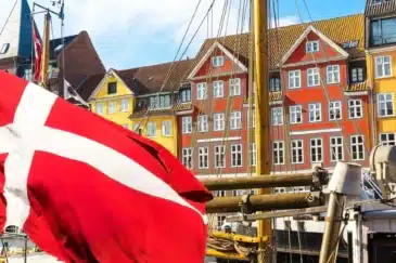 Kuvassa on eloisa ja ikoninen näkymä Kööpenhaminan kuuluisasta Nyhavnin satama-alueesta. Etualalla suuri Tanskan lippu, "Dannebrog", liehuu voimakkaasti tuulessa. Se on kiinnitetty yhteen niistä monista historiallisista puurakenteisista purjealuksista, jotka reunustavat kanavaa. Taustalla on Nyhavnille tunnusomainen rivi värikkäitä, kapeita taloja. Nämä 1600- ja 1700-luvuilta peräisin olevat rakennukset on maalattu kirkkailla keltaisen, oranssin ja punaisen sävyillä, ja niiden valkoiset ikkunankehykset erottuvat selkeästi. Aurinkoinen sää ja sininen taivas täydentävät tämän postikorttimaisen kauniin ja vilkkaan näkymän.