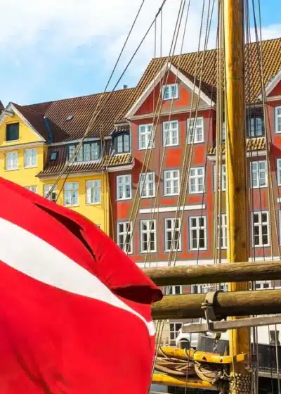 Kuvassa on eloisa ja ikoninen näkymä Kööpenhaminan kuuluisasta Nyhavnin satama-alueesta. Etualalla suuri Tanskan lippu, "Dannebrog", liehuu voimakkaasti tuulessa. Se on kiinnitetty yhteen niistä monista historiallisista puurakenteisista purjealuksista, jotka reunustavat kanavaa. Taustalla on Nyhavnille tunnusomainen rivi värikkäitä, kapeita taloja. Nämä 1600- ja 1700-luvuilta peräisin olevat rakennukset on maalattu kirkkailla keltaisen, oranssin ja punaisen sävyillä, ja niiden valkoiset ikkunankehykset erottuvat selkeästi. Aurinkoinen sää ja sininen taivas täydentävät tämän postikorttimaisen kauniin ja vilkkaan näkymän.