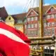 Kuvassa on eloisa ja ikoninen näkymä Kööpenhaminan kuuluisasta Nyhavnin satama-alueesta. Etualalla suuri Tanskan lippu, "Dannebrog", liehuu voimakkaasti tuulessa. Se on kiinnitetty yhteen niistä monista historiallisista puurakenteisista purjealuksista, jotka reunustavat kanavaa. Taustalla on Nyhavnille tunnusomainen rivi värikkäitä, kapeita taloja. Nämä 1600- ja 1700-luvuilta peräisin olevat rakennukset on maalattu kirkkailla keltaisen, oranssin ja punaisen sävyillä, ja niiden valkoiset ikkunankehykset erottuvat selkeästi. Aurinkoinen sää ja sininen taivas täydentävät tämän postikorttimaisen kauniin ja vilkkaan näkymän.