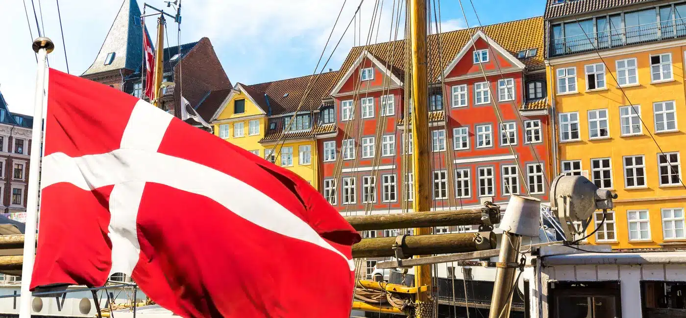 Kuvassa on eloisa ja ikoninen näkymä Kööpenhaminan kuuluisasta Nyhavnin satama-alueesta. Etualalla suuri Tanskan lippu, "Dannebrog", liehuu voimakkaasti tuulessa. Se on kiinnitetty yhteen niistä monista historiallisista puurakenteisista purjealuksista, jotka reunustavat kanavaa. Taustalla on Nyhavnille tunnusomainen rivi värikkäitä, kapeita taloja. Nämä 1600- ja 1700-luvuilta peräisin olevat rakennukset on maalattu kirkkailla keltaisen, oranssin ja punaisen sävyillä, ja niiden valkoiset ikkunankehykset erottuvat selkeästi. Aurinkoinen sää ja sininen taivas täydentävät tämän postikorttimaisen kauniin ja vilkkaan näkymän.