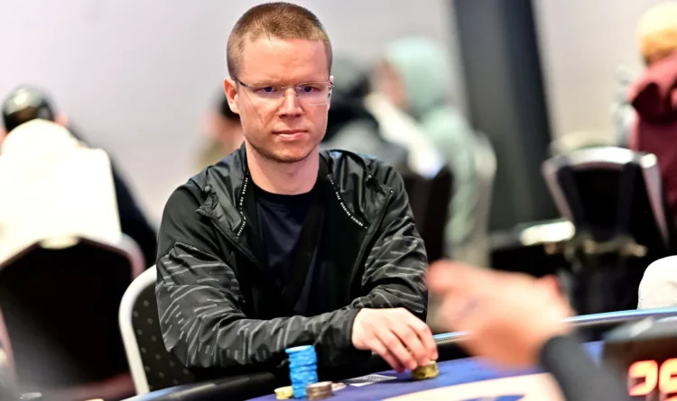 Mies istuu pokeripöydässä. Miehellä on tuima ilme kasvoillaan ja hänellä on päällään tumma paita. Miehen edessä on pelimerkkejä.