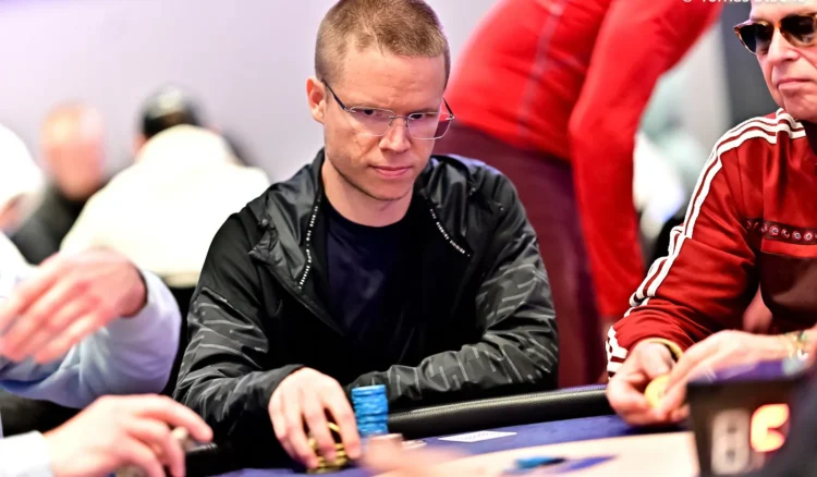 Mies istuu pokeripöydässsä. Hänellä on päällään tumma huppari. Oikealla puolella istuu toinen mies punaisessa verkkatakissa.