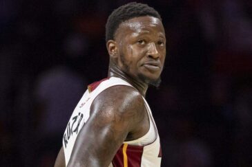 Kuvassa on koripalloilija Terry Rozier kesken Miami Heat -ottelun. Hän seisoo kentällä hieman sivuttain, mutta kääntää päätään ja katsoo taakseen olkansa yli intensiivisellä ja keskittyneellä ilmeellä. Rozierilla on yllään Heatin valkoinen pelipaita, jossa on punakeltaiset yksityiskohdat, ja hänen tatuoidut käsivartensa ovat selvästi näkyvissä. Kuvan tausta on sumennettu, mikä korostaa pelaajaa ja vangitsee pelin keskittyneen hetken.