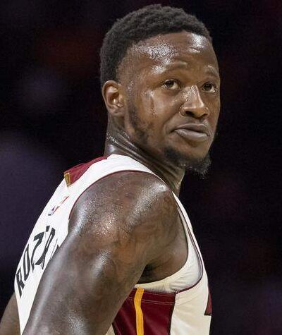 Kuvassa on koripalloilija Terry Rozier kesken Miami Heat -ottelun. Hän seisoo kentällä hieman sivuttain, mutta kääntää päätään ja katsoo taakseen olkansa yli intensiivisellä ja keskittyneellä ilmeellä. Rozierilla on yllään Heatin valkoinen pelipaita, jossa on punakeltaiset yksityiskohdat, ja hänen tatuoidut käsivartensa ovat selvästi näkyvissä. Kuvan tausta on sumennettu, mikä korostaa pelaajaa ja vangitsee pelin keskittyneen hetken.