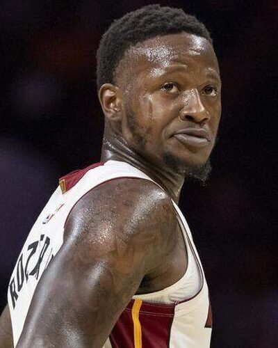 Kuvassa on koripalloilija Terry Rozier kesken Miami Heat -ottelun. Hän seisoo kentällä hieman sivuttain, mutta kääntää päätään ja katsoo taakseen olkansa yli intensiivisellä ja keskittyneellä ilmeellä. Rozierilla on yllään Heatin valkoinen pelipaita, jossa on punakeltaiset yksityiskohdat, ja hänen tatuoidut käsivartensa ovat selvästi näkyvissä. Kuvan tausta on sumennettu, mikä korostaa pelaajaa ja vangitsee pelin keskittyneen hetken.