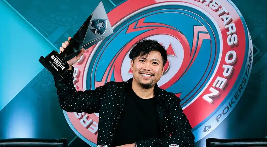 Kuvassa on onnellinen, tummahiuksinen mies, joka juhlii voittoaan PokerStars Open -turnauksessa. Hän hymyilee leveästi kameralle ja pitelee ylpeänä koholla modernia, terävänmuotoista pokaalia, jossa lukee "MANCHESTER". Voittaja istuu pokeripöydän ääressä, ja hänen edessään on järjestelmällisissä pinoissa suuri määrä vihreitä ja valkoisia pelimerkkejä. Merkkien eteen on asetettu näytille hänen voittokätensä, ässäpari. Miehellä on yllään musta paita ja tyylikäs musta, valkopilkullinen takki. Taustalla loistaa suuri, punasinivalkoinen "POKERSTARS OPEN" -tapahtuman logo. Kuva on täynnä aitoa voitonriemua.